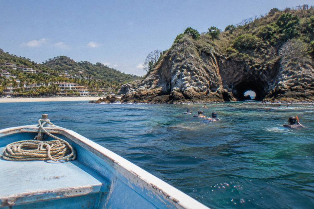 A small boat near a rocky island shoreline with snorkelers over a coral reef in clear water