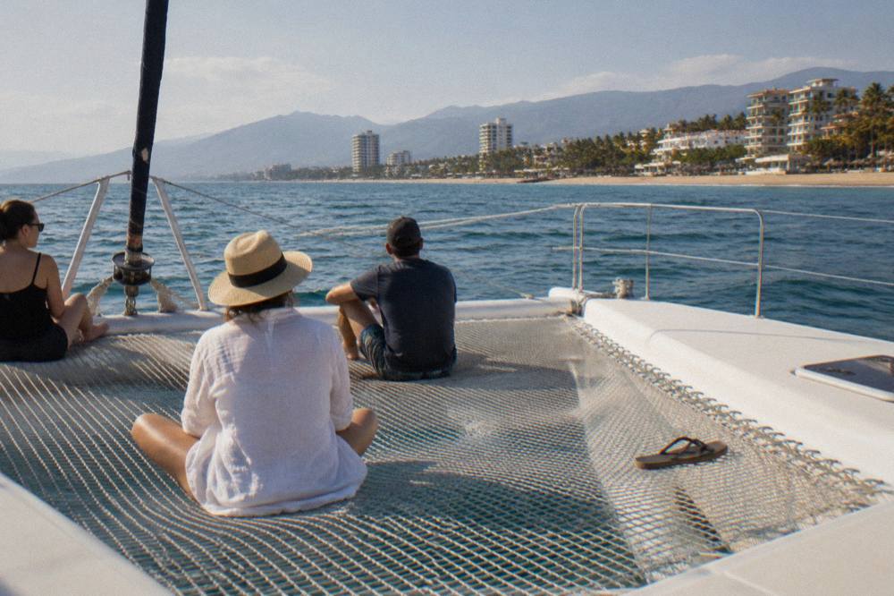 A wide catamaran deck with netting at the bow and passengers seated along the trampoline in sunlight