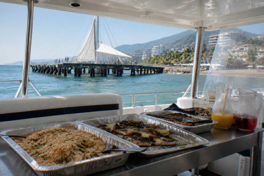 A buffet lunch laid out on a catamaran deck table with trays of food and drinks under a hardtop