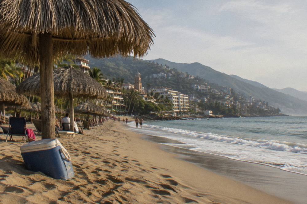 Long sandy town beach with palapa umbrellas, gentle surf and hills along the shoreline