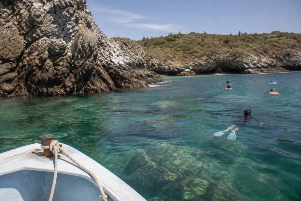 A small boat anchored near a rocky island shoreline with snorkelers in clear water