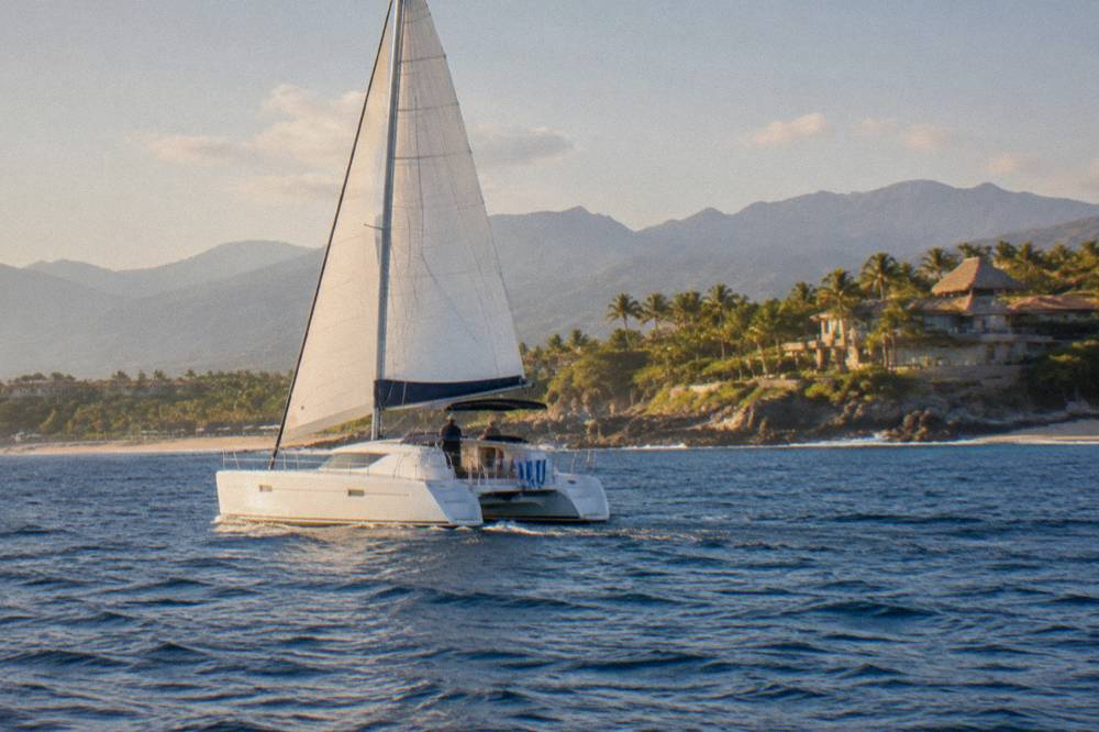 A catamaran under sail on open blue water with hilly coastline in the distance