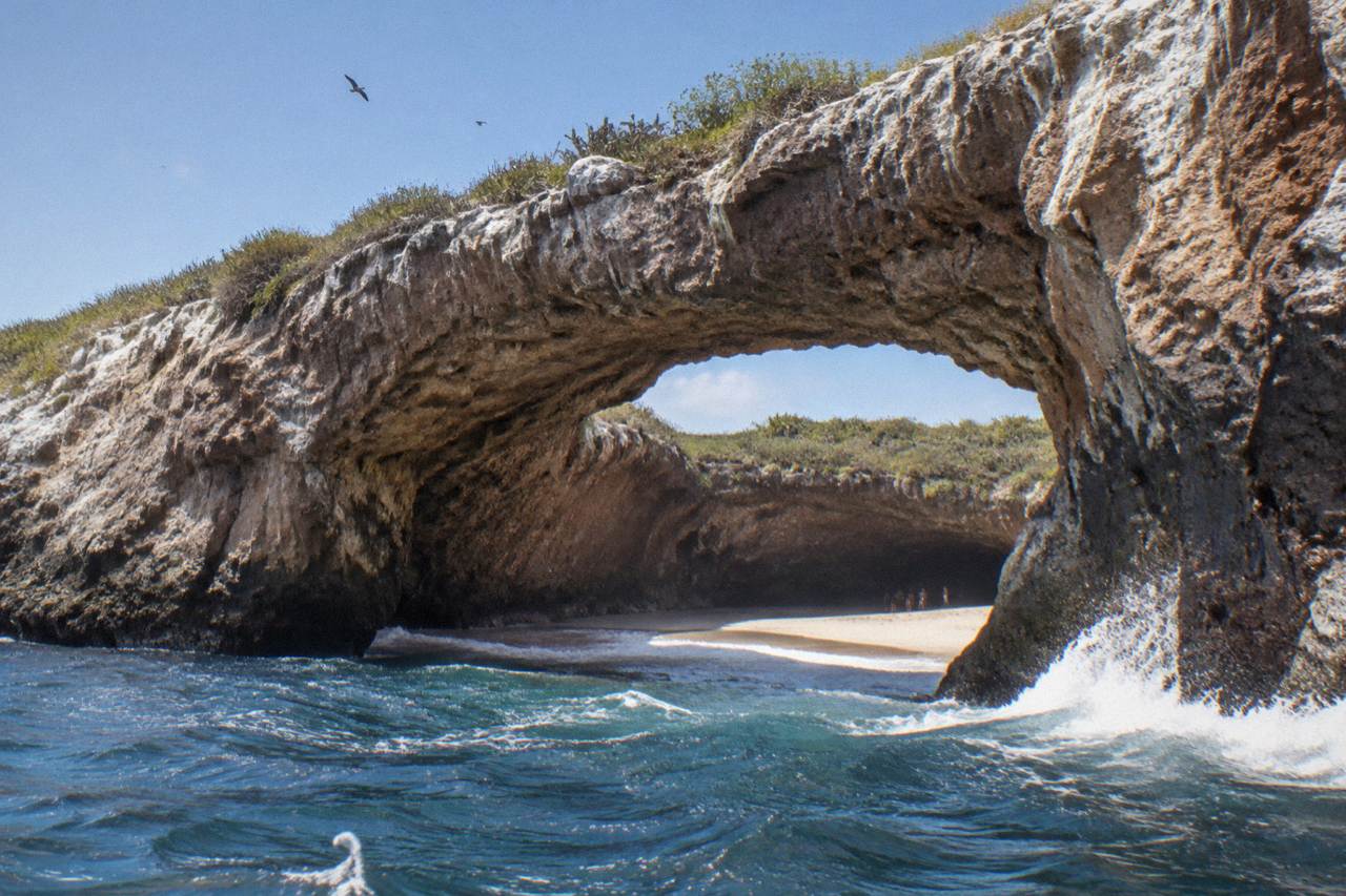 A stone arch above a secluded sandy cove surrounded by cliffs on a small Pacific island