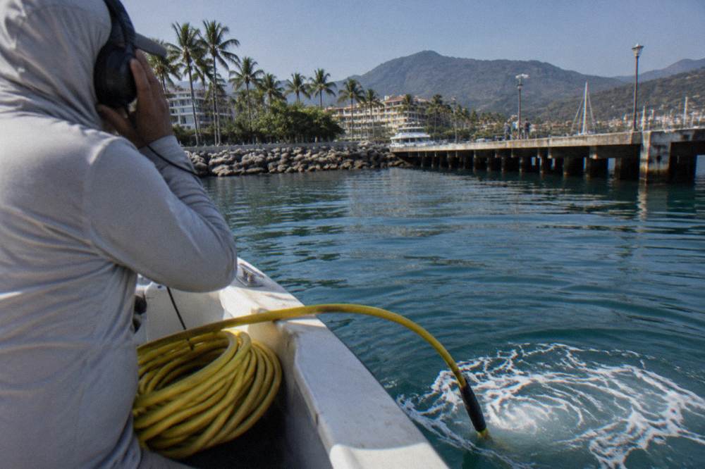 A hydrophone cable trailing into blue water from the side of a boat while a crew member holds the listening headset
