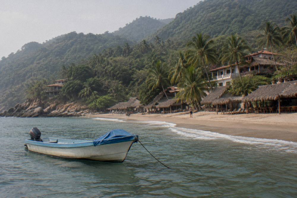 A small boat anchored near a sandy beach with palm-fringed hills behind