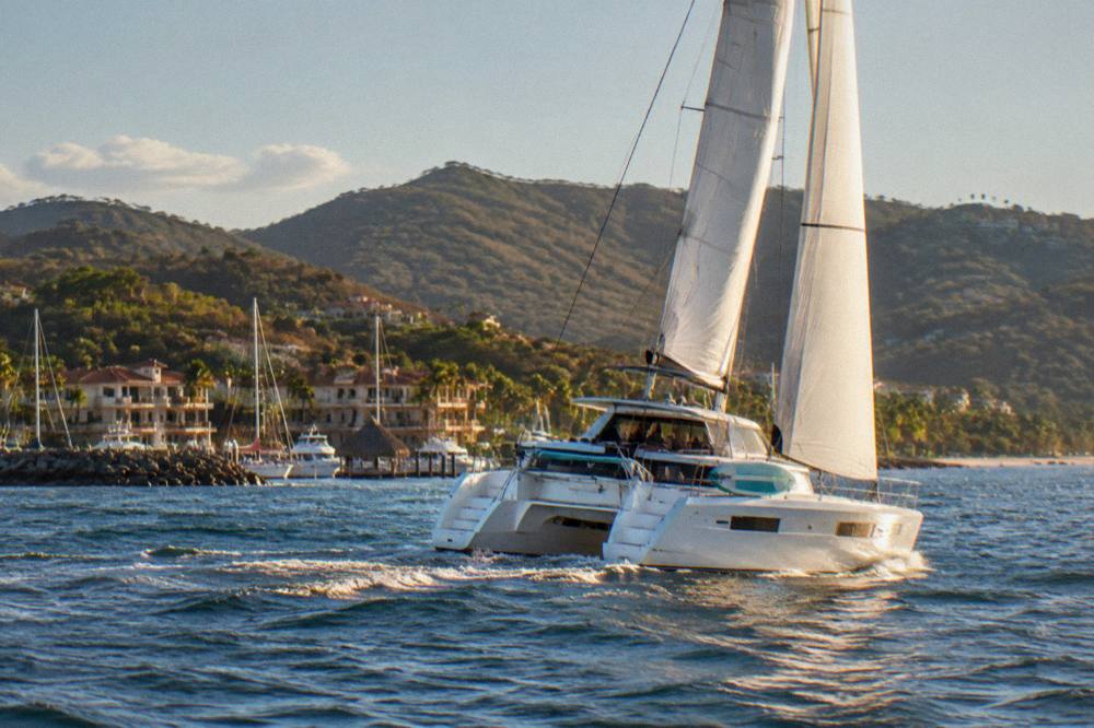 Catamaran under sail on open water with a hilly shoreline behind