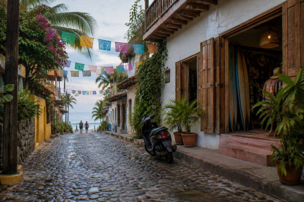 Cobblestone street with colorful flags leading toward a beach town shoreline in morning light