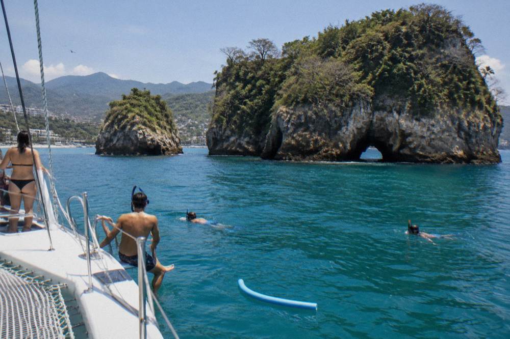 A catamaran anchored near rocky islands in Banderas Bay with snorkelers preparing to enter clear water