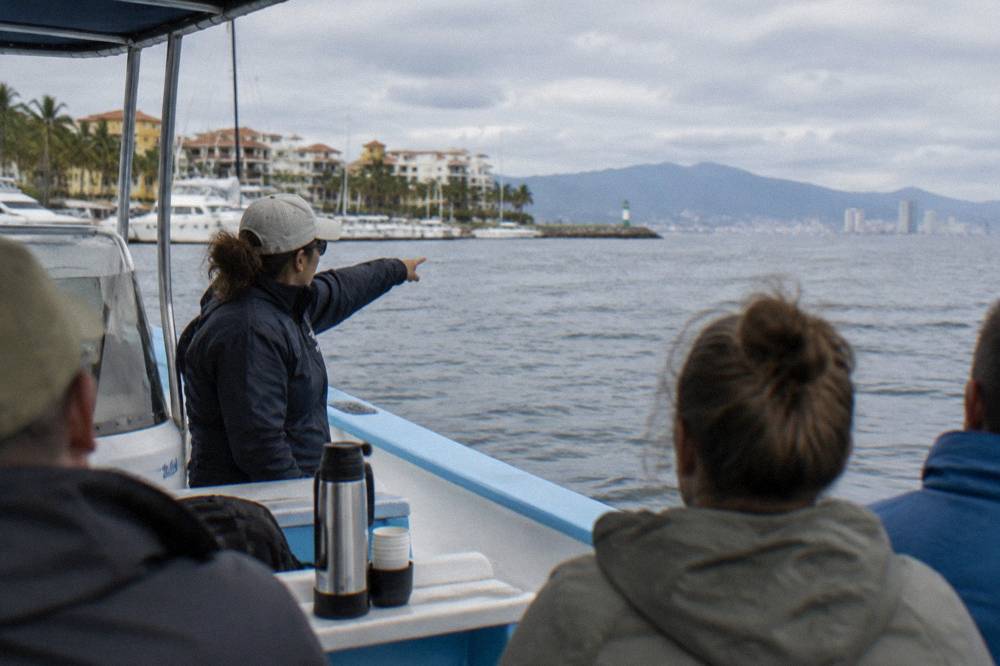 A marine biologist pointing toward open water while speaking to passengers on a small whale watching boat